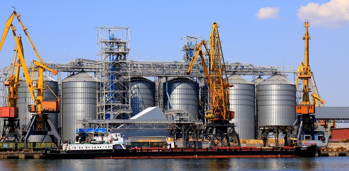 Harbor cranes unload barge at Odesa Seaport amid large metal tanks with wheat, grain, food. 