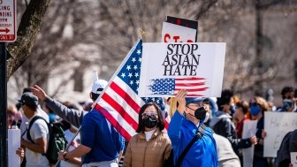 A man stands in a group of protestors wearing a masks, holding a sign with an American flag on it that says Stop Asian Hate.