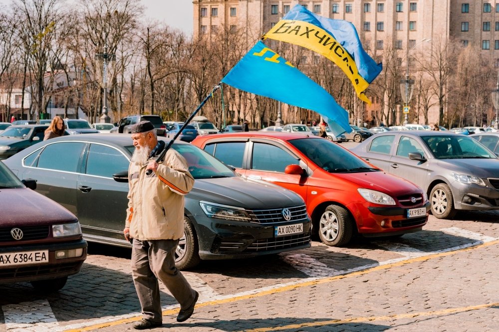 Crimean Tatar man with big beard and traditional cap walks on the city's street with Crimea Tatars and Ukrainian flags. 