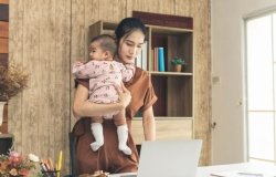 Mother stand and working online with a laptop holding her baby daughter at home office. 